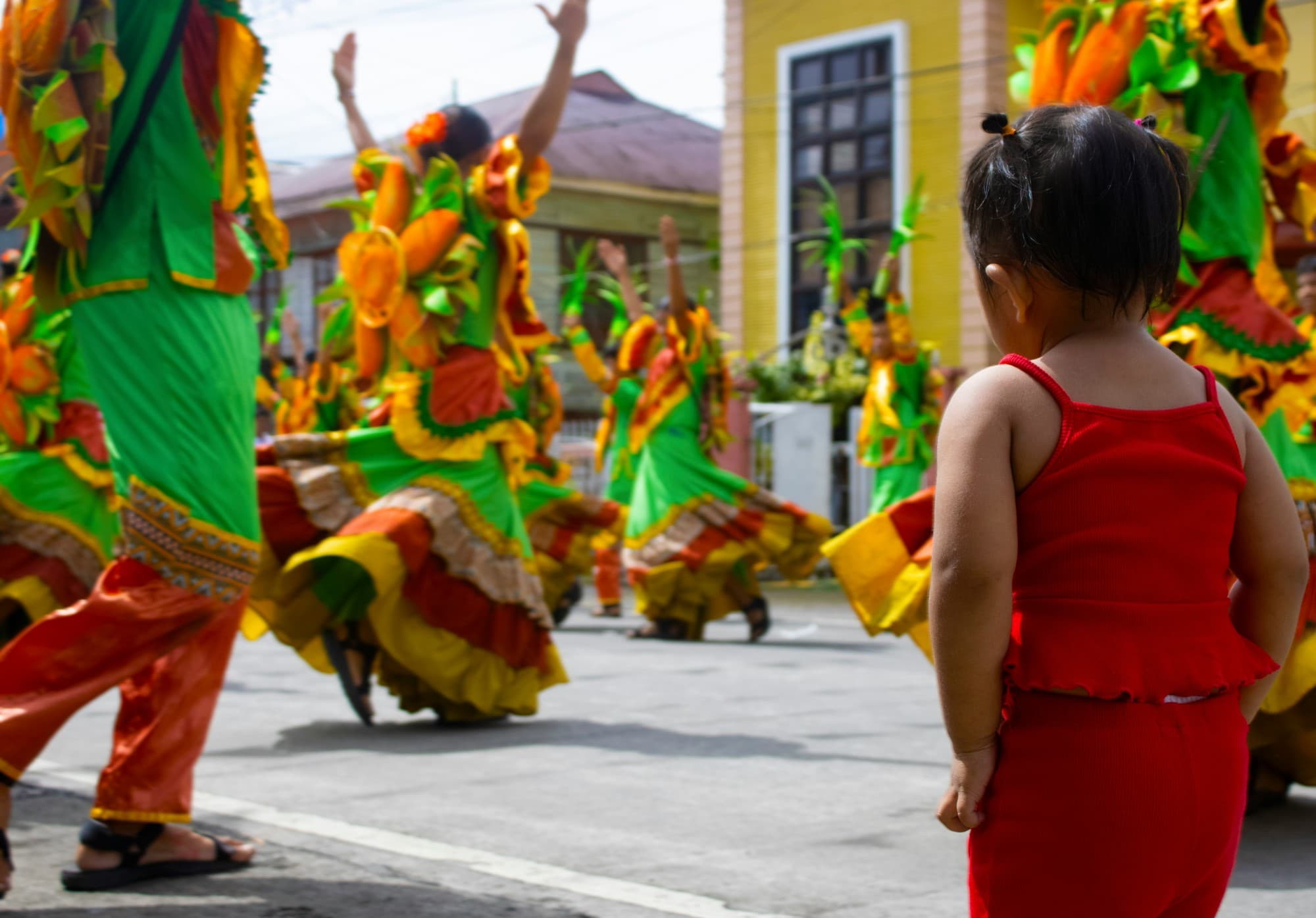 A small girl in red watching dancers in green, orange and yellow costumes at a street festival